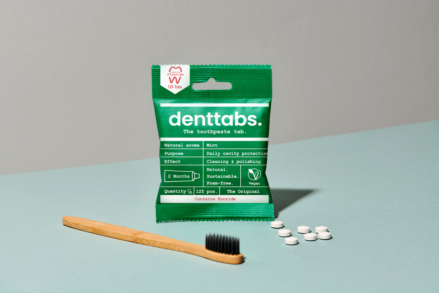 Packaging of denttabs with a wooden toothbrush and white tablets on a light blue surface.