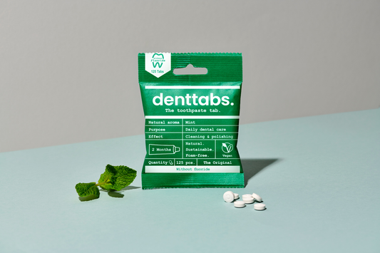 Green package of denttabs toothpaste tabs on a light blue surface with mint leaves.